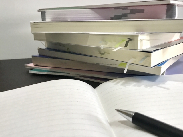 山積みに積み上げられた何冊もの参考書や問題集と、手前にある白紙のノートに置かれたシャープペンシル。夏休みの計画倒れの焦りを表すイメージ風景